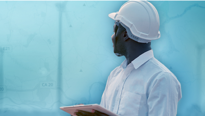 A man with a white hard hat, holding a tablet while looking out onto a blue background 