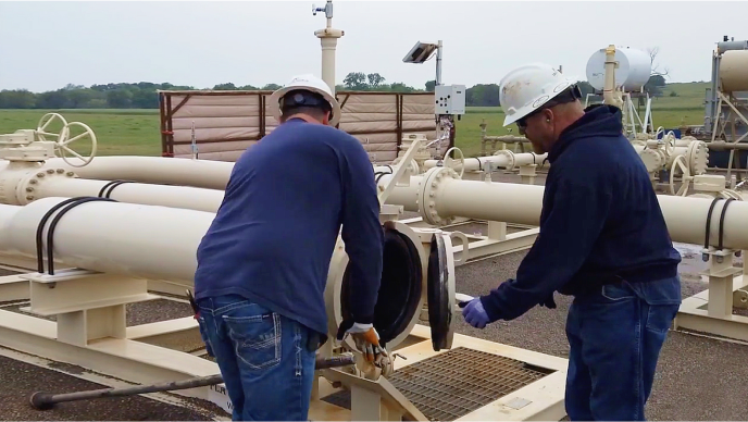 Two mobile workers in white hard hats performing a pipeline inspection outdoors 