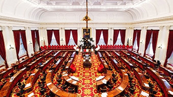 A large room with many wood tables, red window drapes, and a large chandelier representing a congressional room