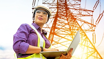 A female construction worker wearing a yellow vest with an open laptop computer in front of a utility tower