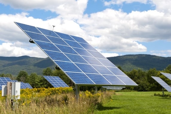 A photo of a large shining blue solar panel in a sunlit green field with a cloudy blue sky overhead