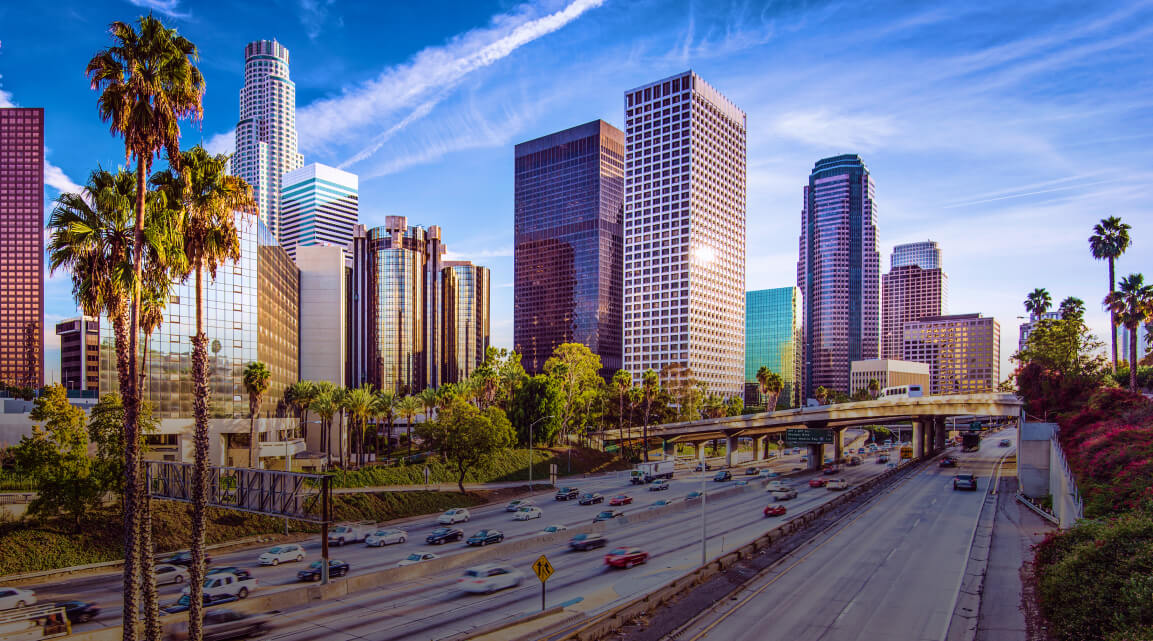 Downtown Los Angeles skyscrapers against a vibrant blue sky with palm trees and a freeway in the foreground