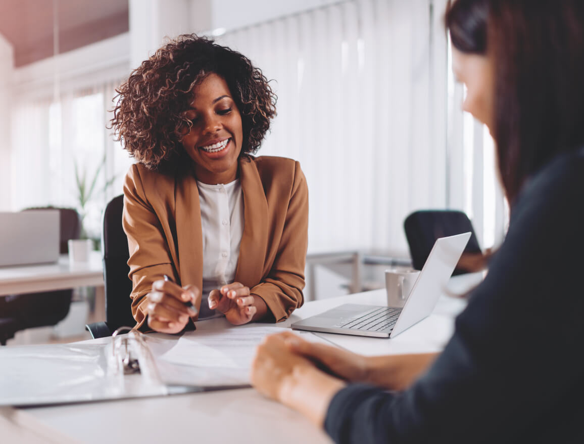 A smiling person wearing a tan blazer speaking to another person across a desk in an office