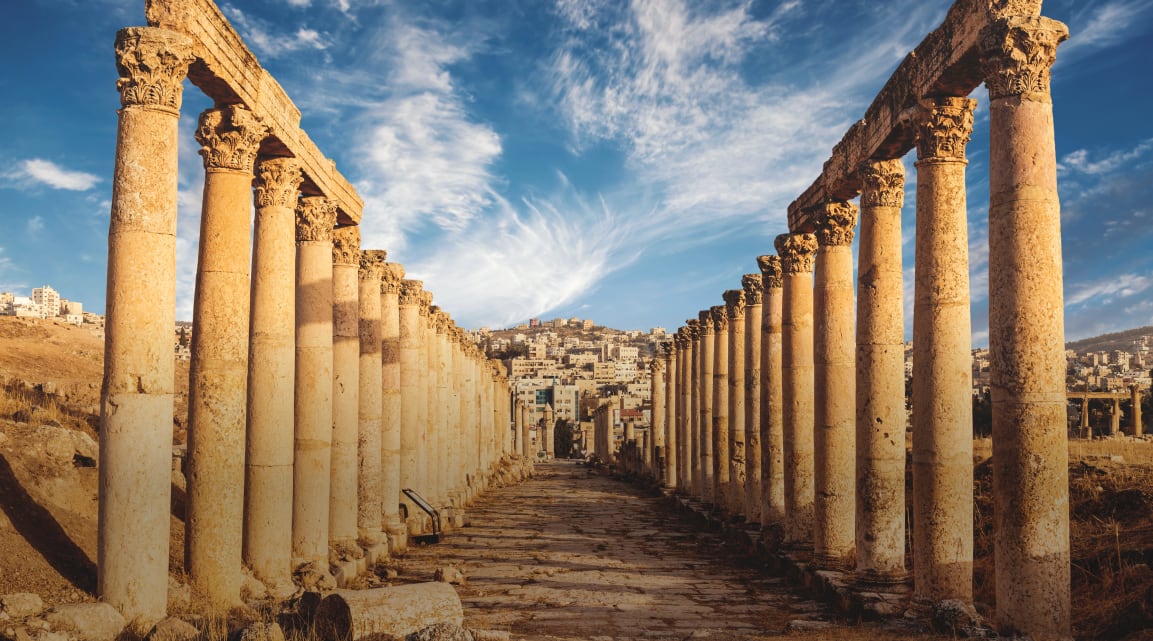 Two rows of ancient columns leading towards a city built on a hill against a cloud-streaked blue sky
