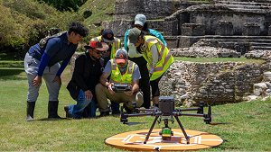 Six researchers gather around a controller and a drone on a landing pad with ancient ruins in the background