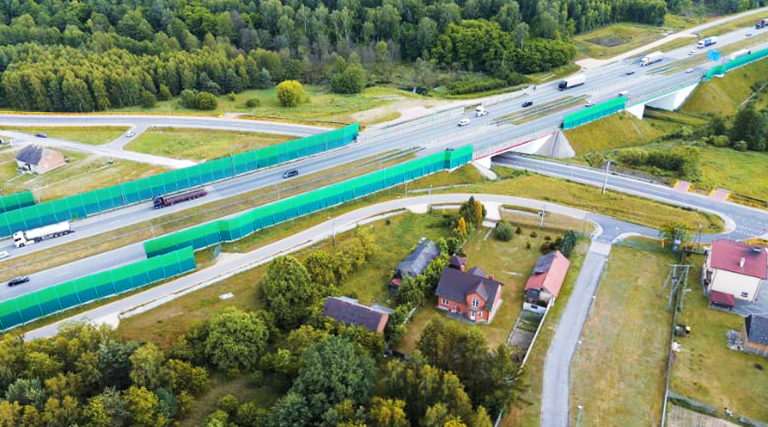 Aerial view of a highway with green sound barriers, an overpass, and nearby houses surrounded by trees.