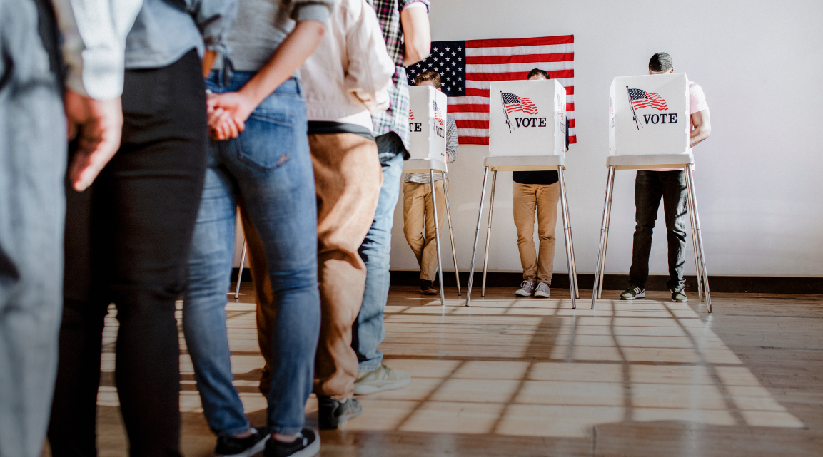 A polling location with 3 people standing behind privacy barriers casting their ballots in front of an American flag
