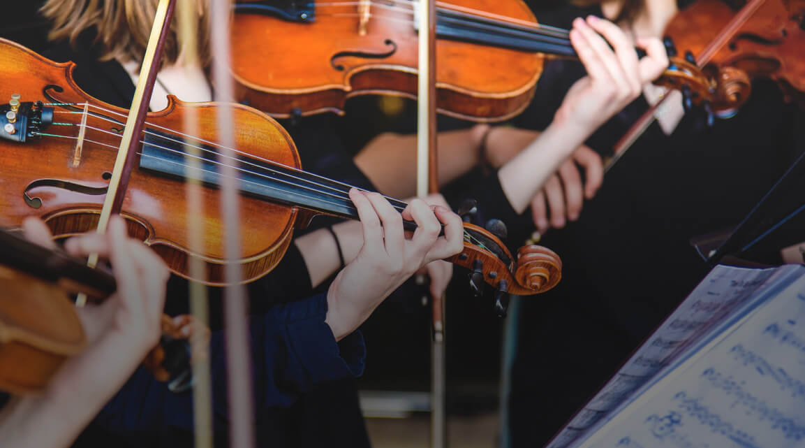 A group of musicians dressed in black playing richly colored wooden violins with sheet music visible in the lower right corner