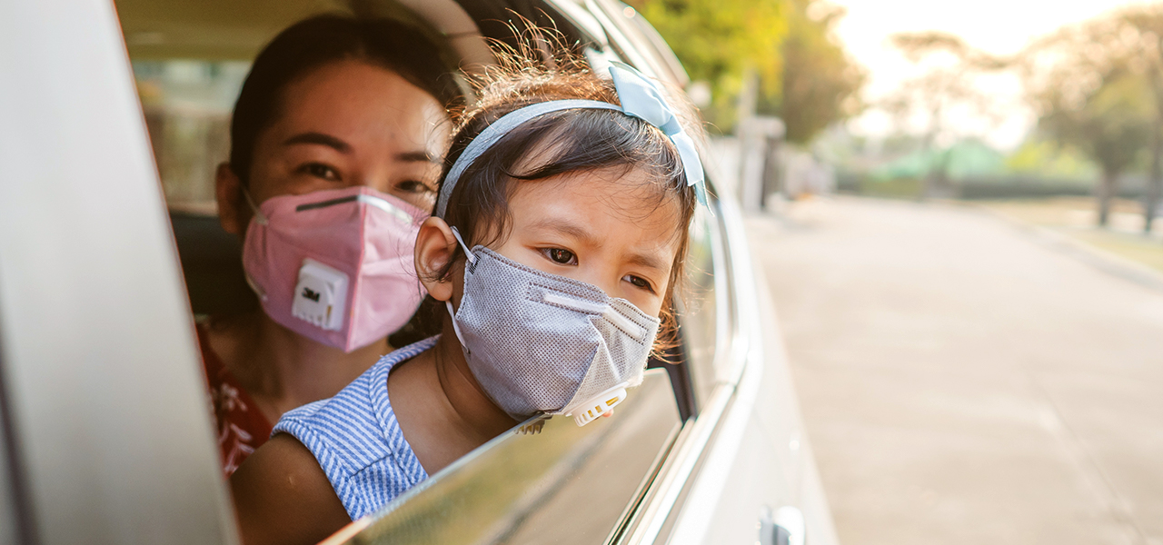 A woman in a car wearing a mask and holding a young child also wearing a mask