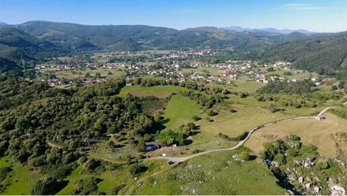 An aerial view of a village with red-roofed houses in a green valley, surrounded by hills and forested mountains under a clear blue sky