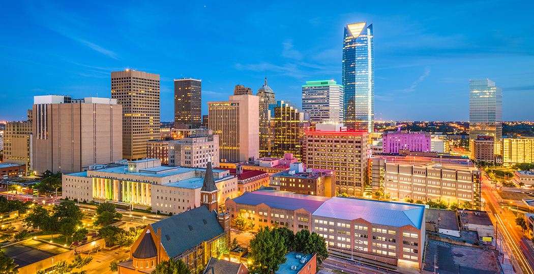 An aerial view of a colorful city full of skyscrapers brightly lit for twilight under a deep blue sky