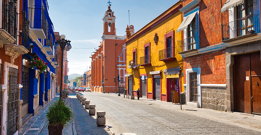 A street in Mexico lined with colorful, historic buildings under a blue sky overlaid with a play button