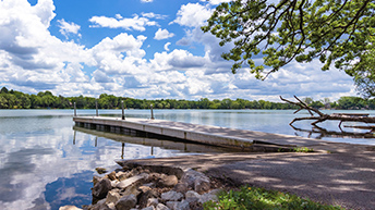 A shaded boat pier in a placid lake under a blue sky with puffy, white clouds