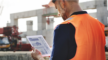 A person in an orange safety vest stands in a sunlit constructions site reviewing plans on a handheld tablet