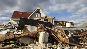 A large pile of rubble sitting in the front yard of the remains of a large white home with a red roof under a cloudy sky