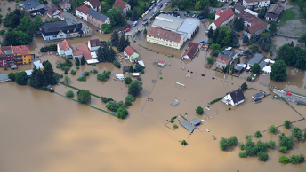 An aerial photo of a suburban neighborhood partially submerged in pale brown floodwater with roofs and treetops visible above the surface