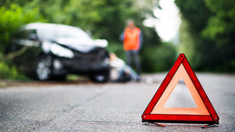A ground-level photo of an orange traffic cone resting on wet road pavement with a crumpled car and bystanders in the background