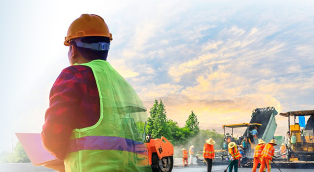 A person in a yellow safety vest and hard hat holding a laptop and looking away into the sunset, with a crew in safety vests repairing a road with heavy equipment in the background