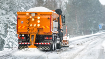 An orange snowblower at work on a snow-covered highway lined with thick growths of tall pine trees