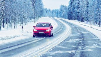 A red sedan driving on an icy winter highway lined with snow-covered pine trees under a hazy white sky