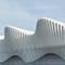 The side of a modern train station with walls in the shape of massive flowing white waves beneath a hazy blue sky