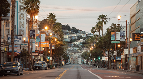 A ground-level view of a San Francisco street lined with shops and palm trees, with terraces of homes and apartment buildings climbing a hillside in the background