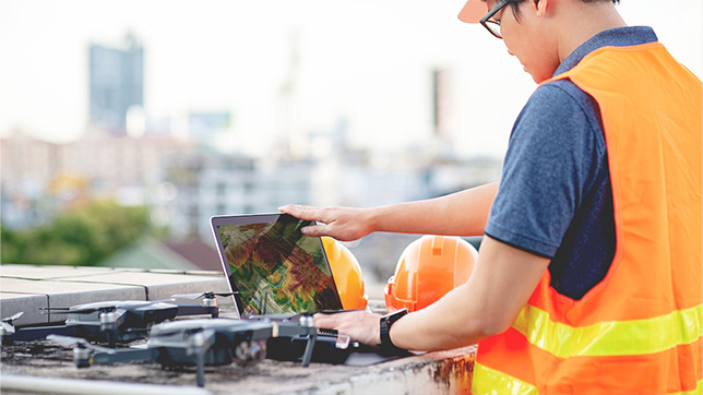 Worker in a safety vest and hard hat looks at a laptop screen, with several small drones parked alongside