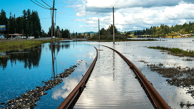 A flooded railway bed under a partly cloudy sky, with water reflecting clouds and nearby utility poles