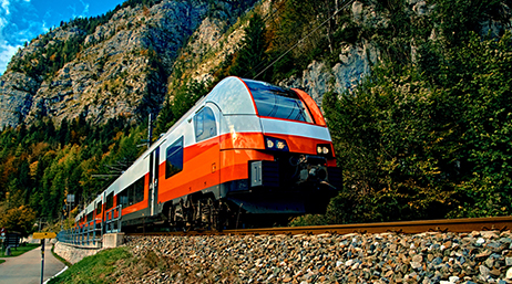 An orange and silver passenger rail moving in front of a forested mountain