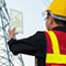 A person in a safety vest and hard hat standing in front of a utility tower and holding a tablet displaying map data