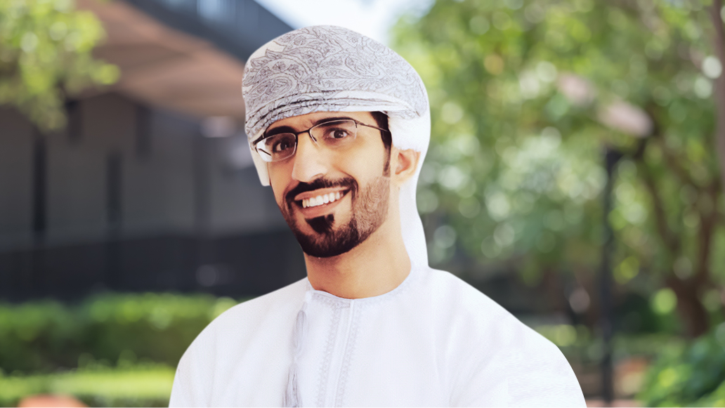 Hussam Al Jabri smiling and wearing a white tunic standing in front of lush greenery in soft focus