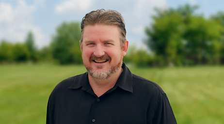 Glen Cook wearing a black collared shirt, smiling and standing outside