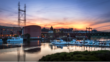 A business complex lit up for nighttime beside a still dock full of white boats against an orange and dark blue sunset sky