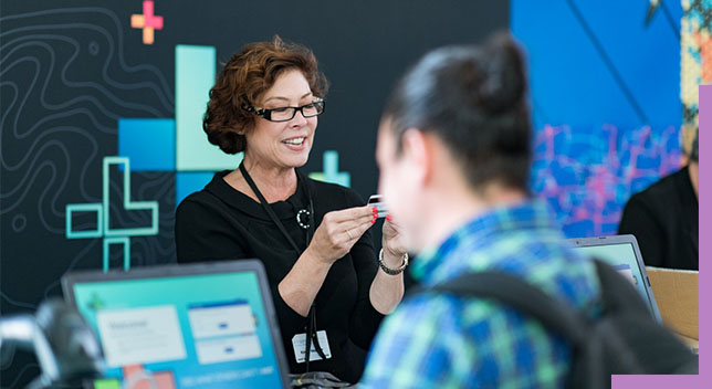 A photo of a conference staffer helping a registrant, while a person in a backpack stands in the foreground