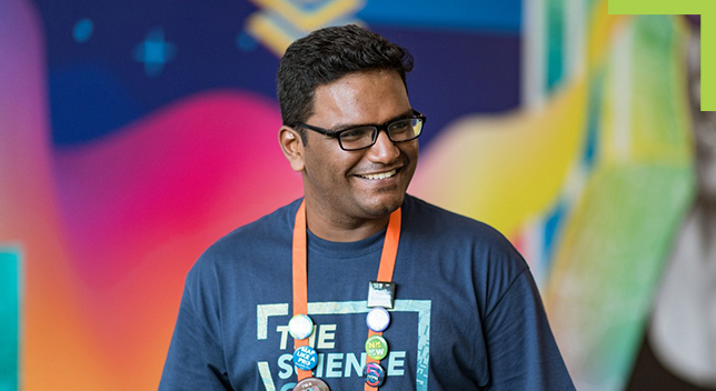 A person smiling wearing an Esri lanyard with pins and a “Science of Where” t-shirt