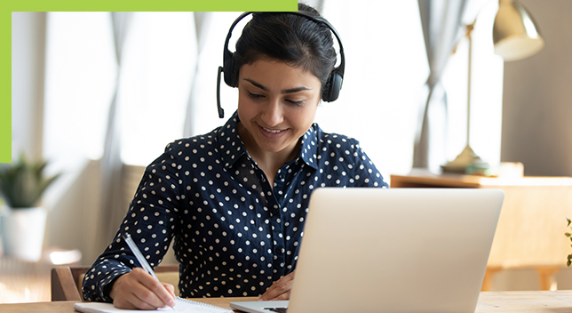 Person mit Headset, die an einem Schreibtisch mit einem geöffneten Laptop sitzt und etwas auf Papier notiert