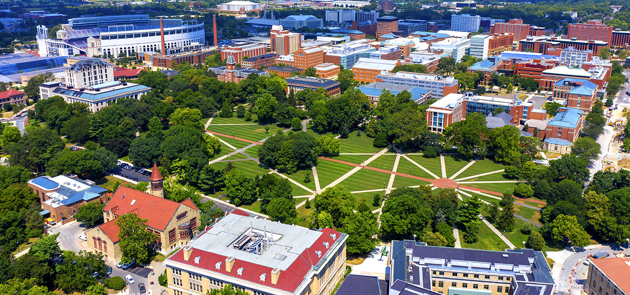 An aerial photo of a university with buildings in blues and reds with a large green tree-filled park in the middle