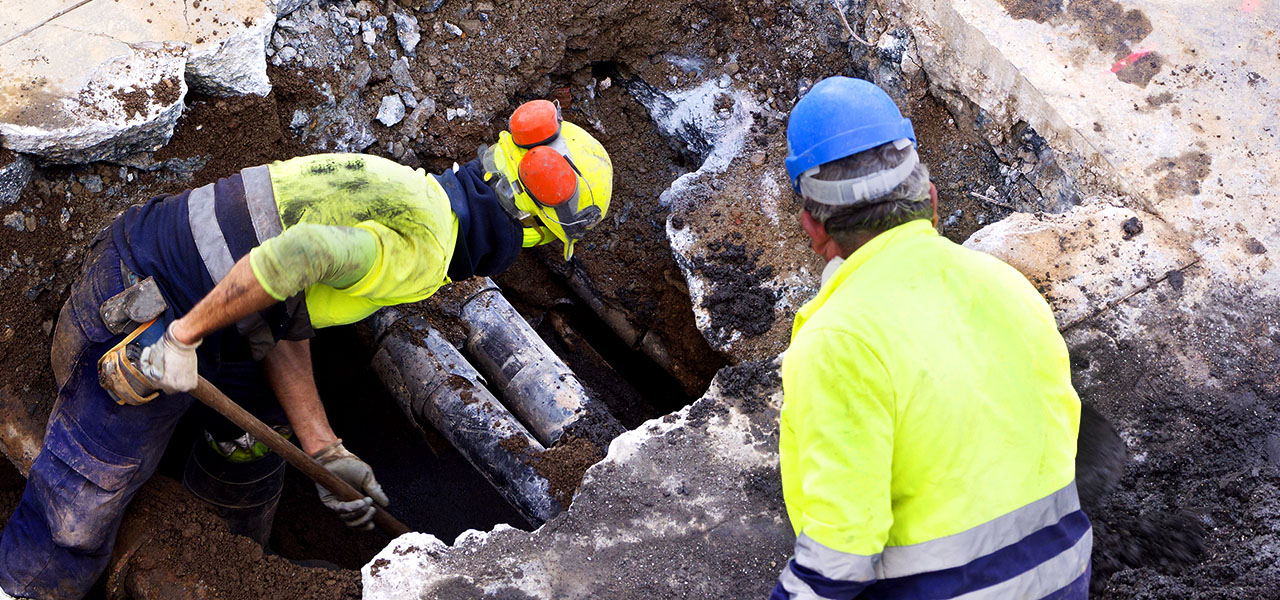 Deux professionnels du terrain en gilet jaune et casque de chantier examinant un gros trou creusé dans un trottoir pour mettre à jour des tuyaux enterrés