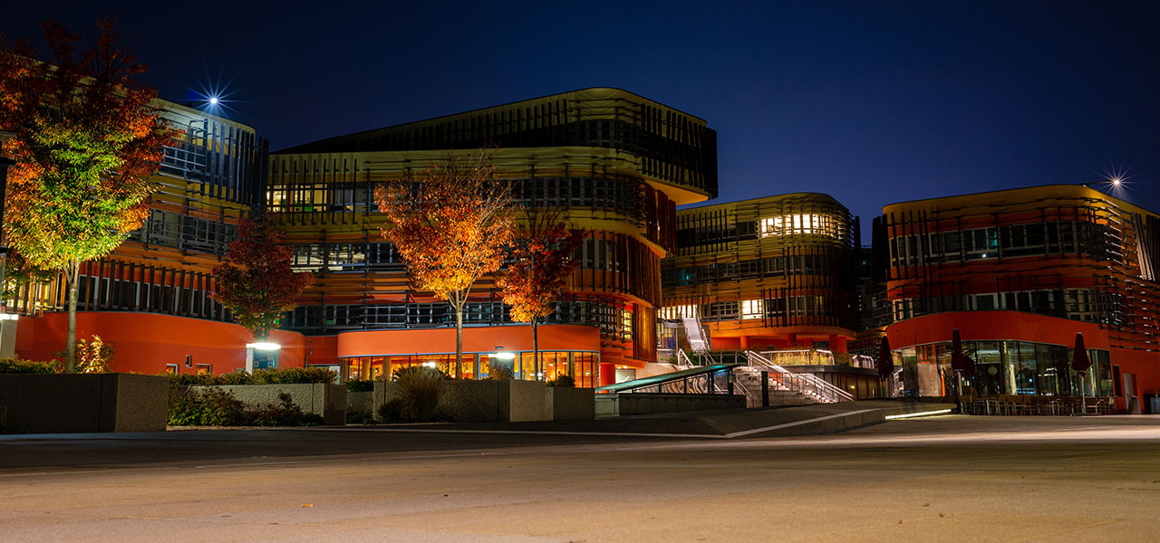 Zdjęcie dużego, dobrze oświetlonego kampusu uniwersyteckiego w kolorach pomarańczowym i żółtym na tle ciemnoniebieskiego nocnego nieba