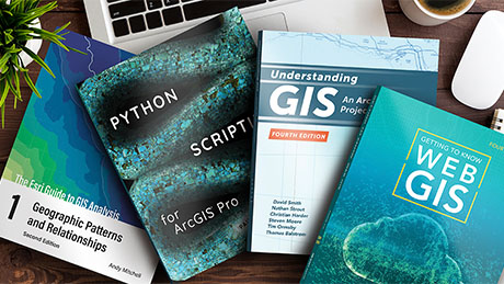A photo of four blue and green Esri Press books and a laptop scattered across an office desk