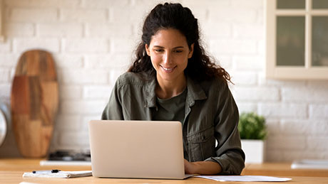 A casually dressed person works at a laptop in a softly lit home setting in shades of white and light brown
