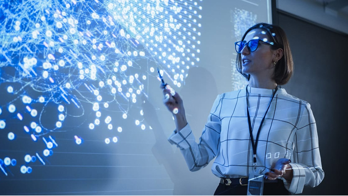 A person wearing glasses and a lanyard pointing to a map projected on a wall overlaid with a play button