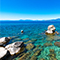 A rocky shoreline with vivid clear blue water dotted with white boulders and rolling blue mountains in the distance