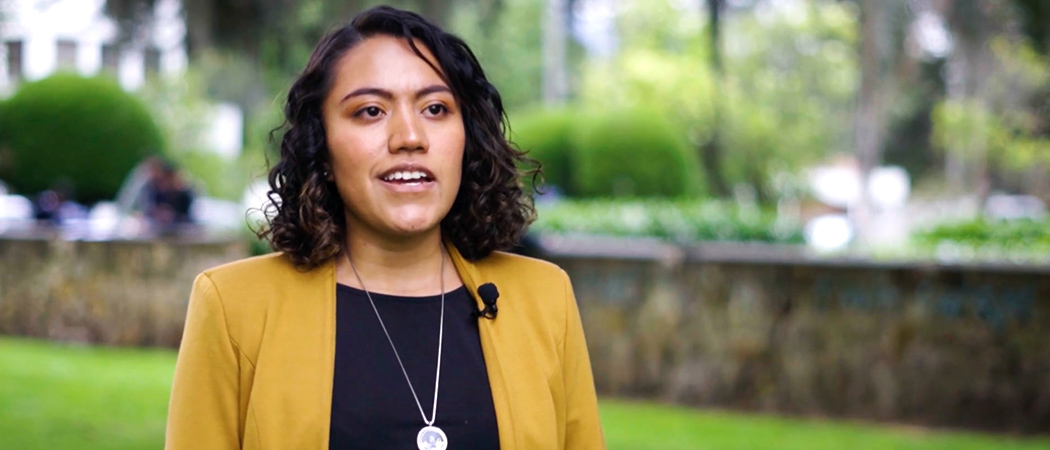 Diana Beltrán wearing a yellow blazer and standing in an outdoor area with a lot of greenery overlaid with a play button