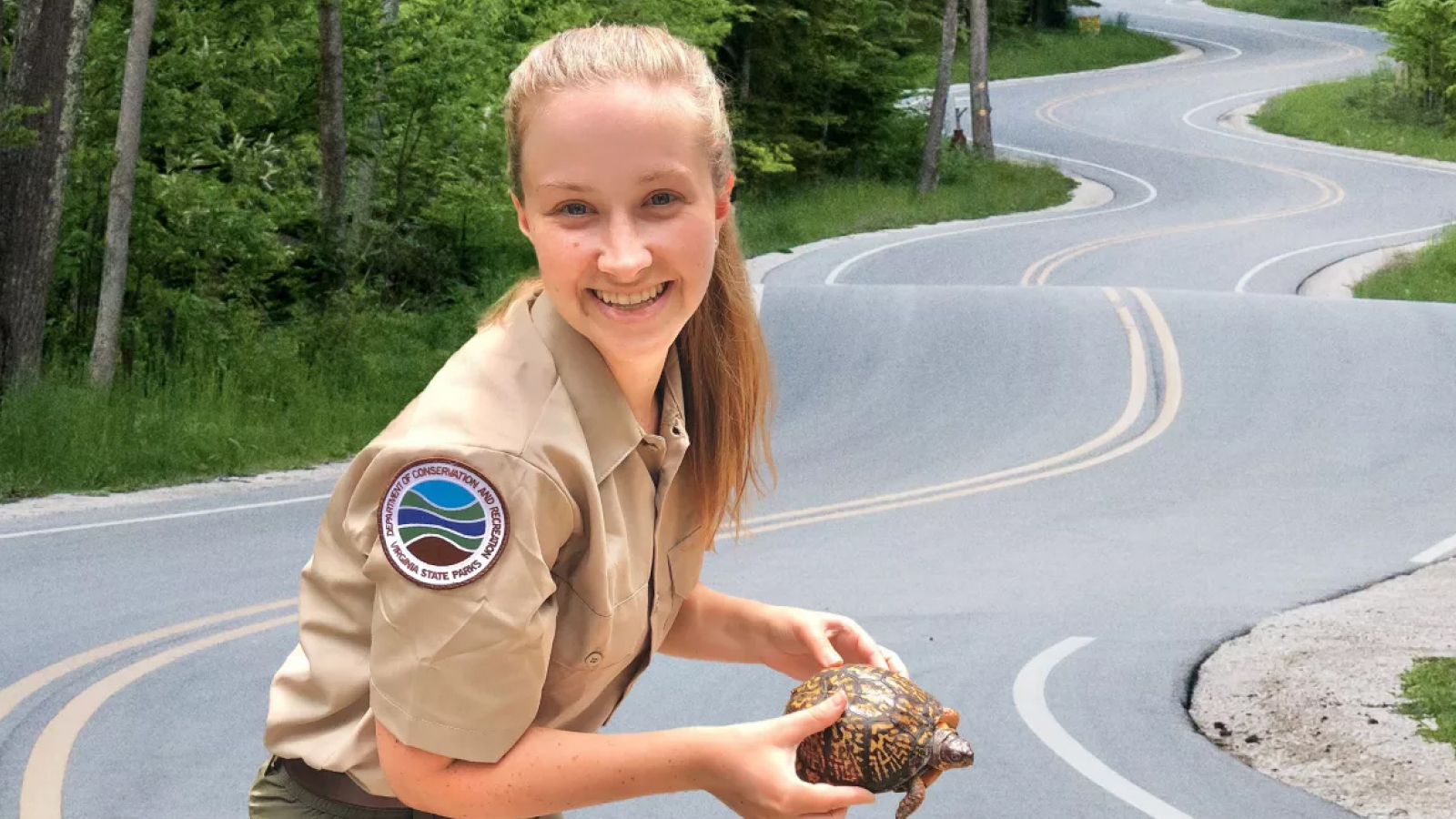  A Virginia State Park employee crouches on a tree-lined road, holding a small tortoise and smiling for the camera