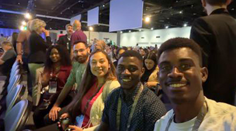 A row of smiling student assistants wearing conference lanyards as they sit in a large plenary audience