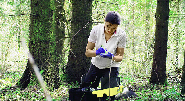 A photo of a casually dressed person kneeling in a forest making notes on a mobile device with a small array of equipment scattered on the ground