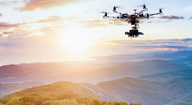 An aerial photo of a drone in flight over a vista of green forest-covered mountains under a bright sunset with blue and pink clouds