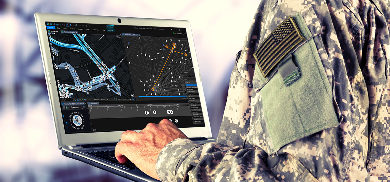 A closeup photo of the hands of a uniformed US soldier in fatigues, typing on a small laptop which displays two maps