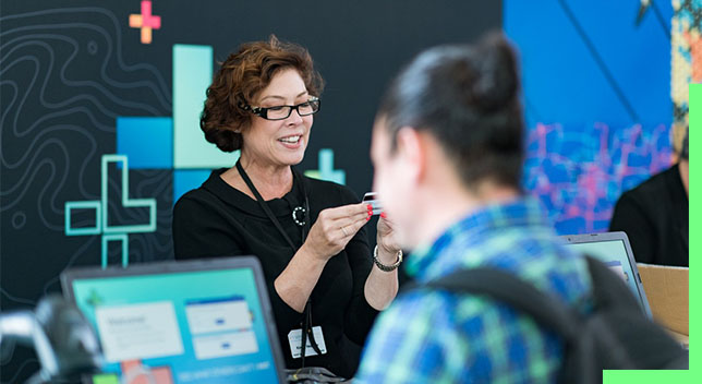 A person looking at an identification card at an event booth with a person in the foreground accessing a computer